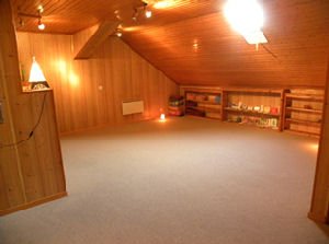 La salle mansardée tout en bois, vide, éclairée par des spots, avec des étagères au fond